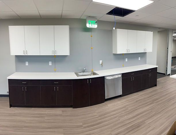 Office breakroom with white countertops, dark cabinets, a sink, and a dishwasher. White upper cabinets. Light wood flooring.