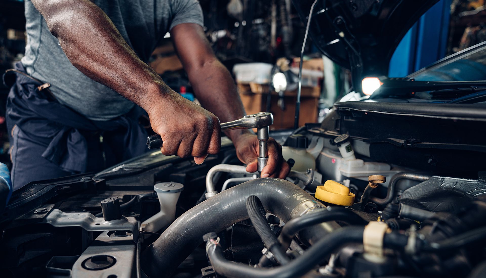 Mechanic using a wrench to work on a car engine under the hood in a garage.