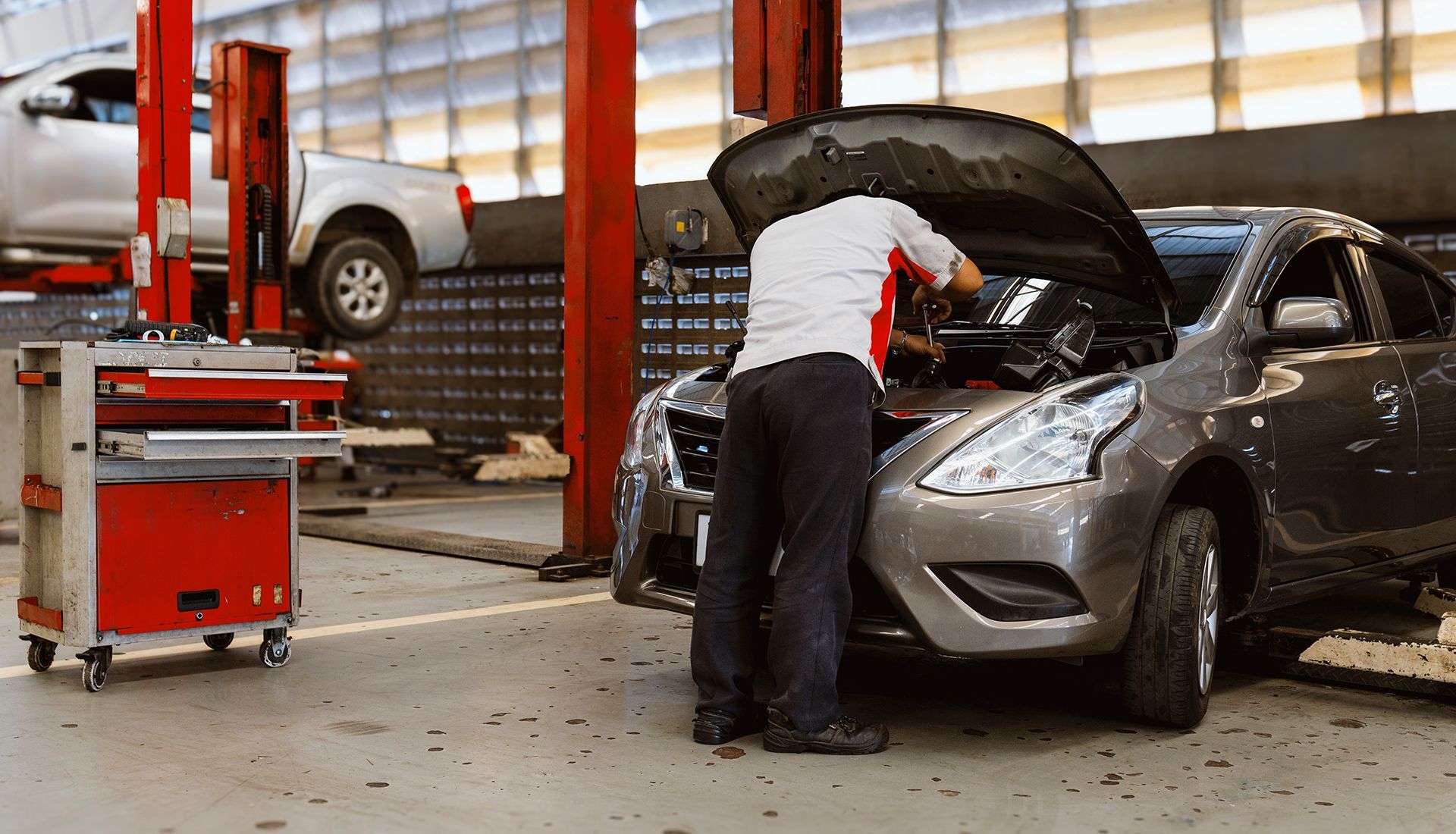 Mechanic working on car engine in a garage. Red lift posts, tool cabinet, gray car.