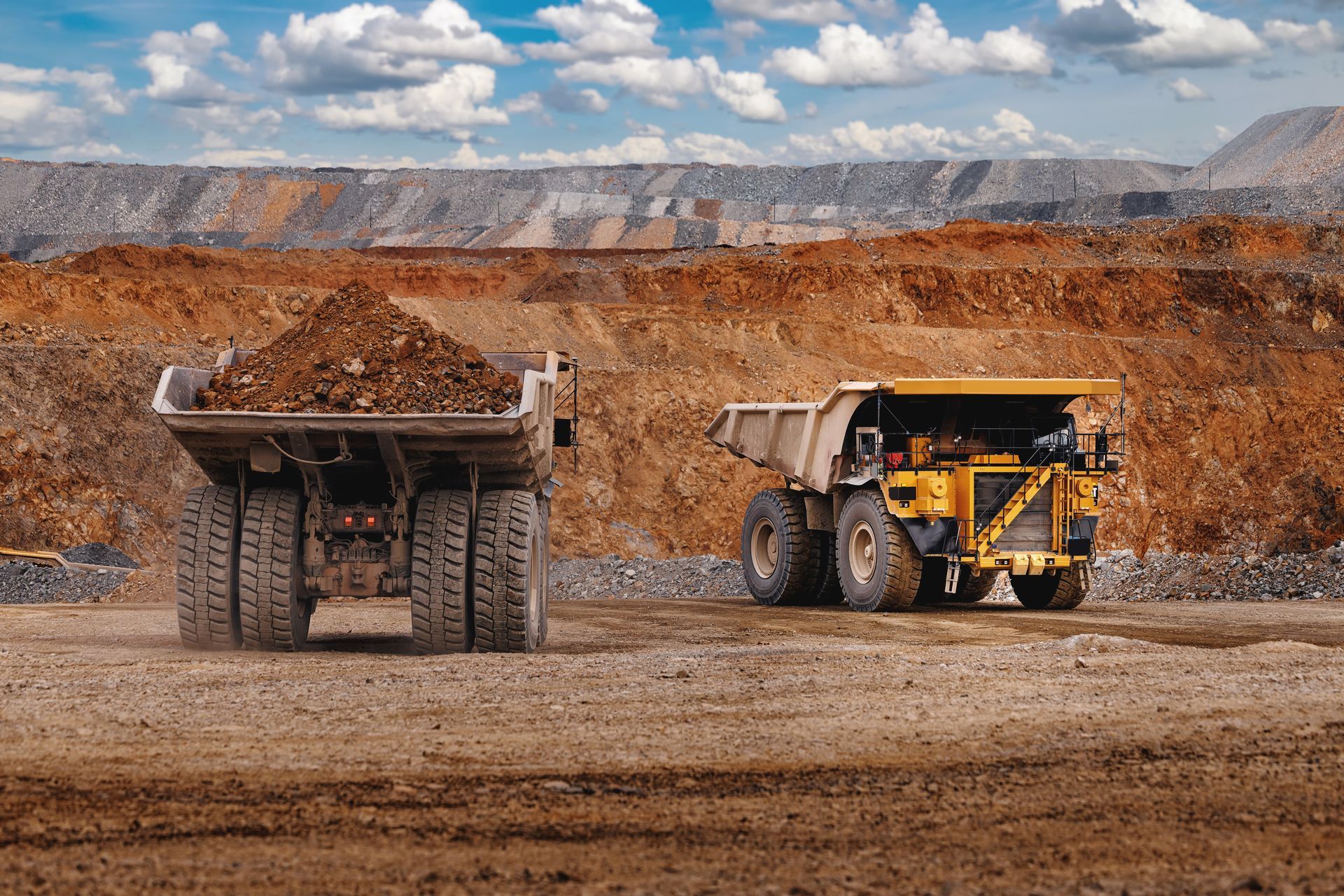 Two large mining dump trucks on a dirt site, one loaded with ore, under a cloudy sky.