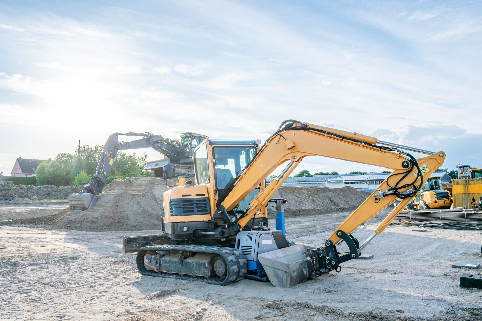 Yellow excavator on a gravel construction site, scooping dirt with a blue truck nearby.