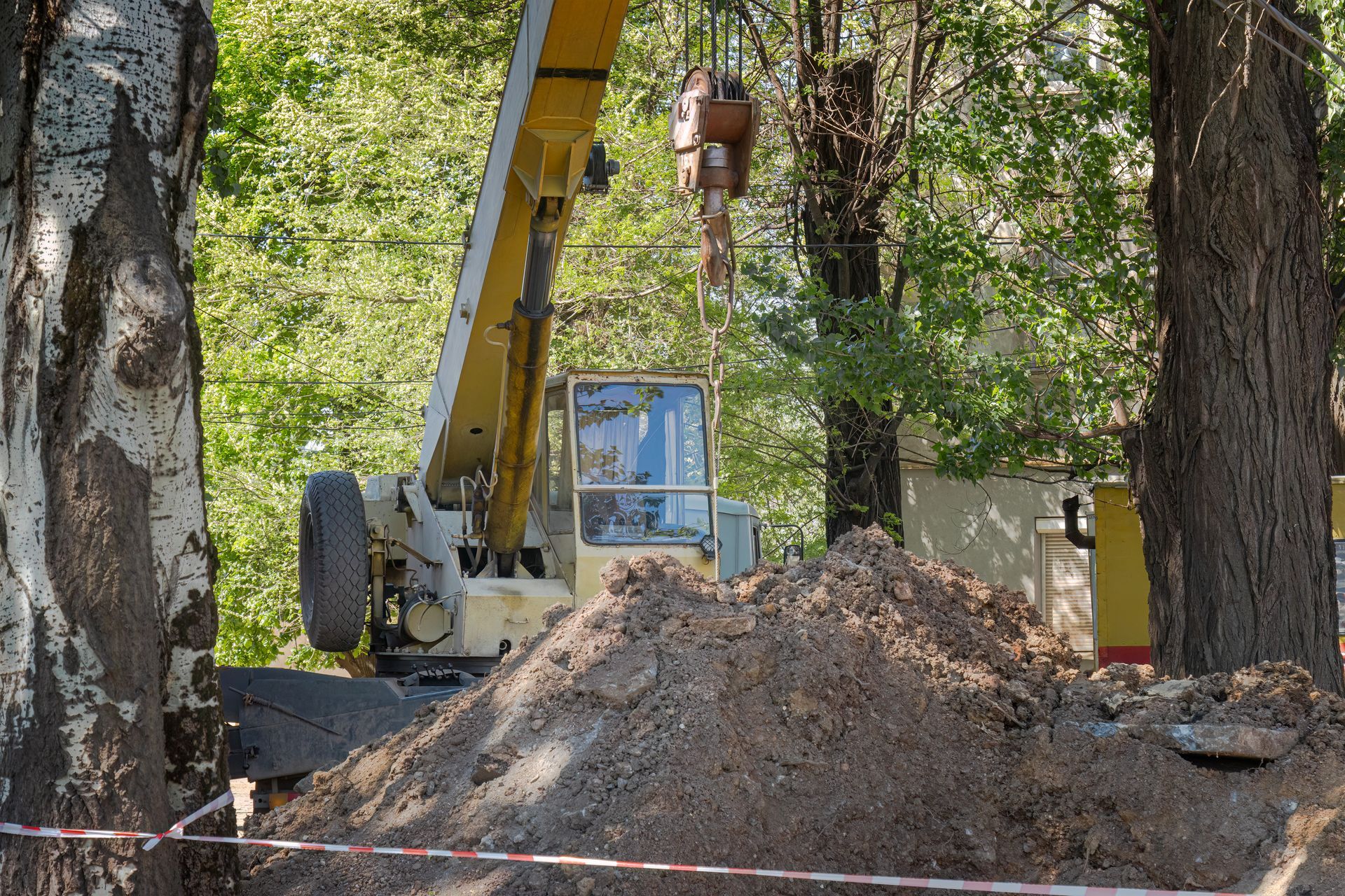 Construction equipment beside a dirt mound among trees, with a worker in a yellow excavator cab