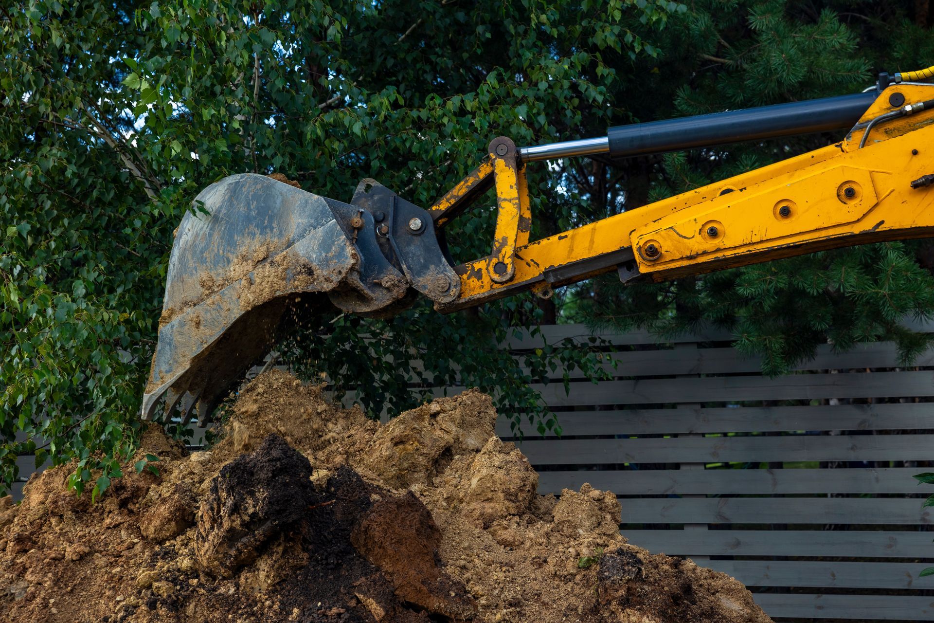 Yellow excavator bucket digging soil beside a wooden fence and green foliage