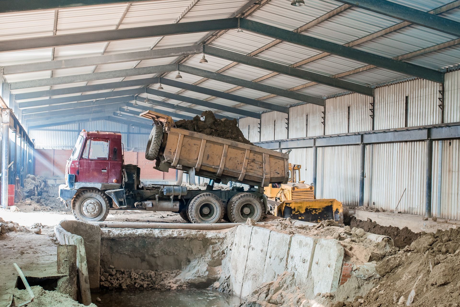 Dump truck in a large warehouse, backing over a concrete pit under construction.