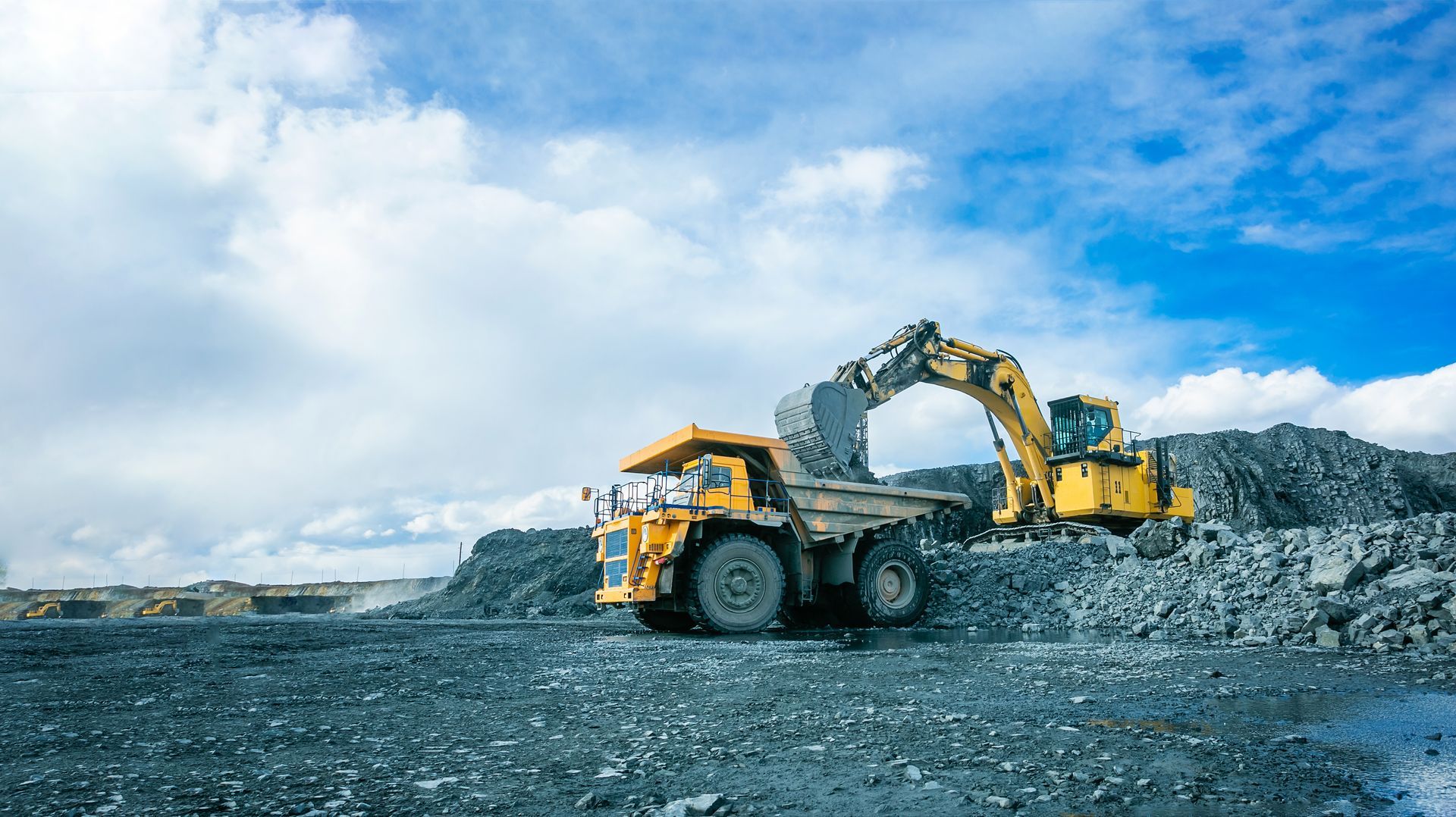 Yellow excavator loading rocks at a construction site under a cloudy blue sky