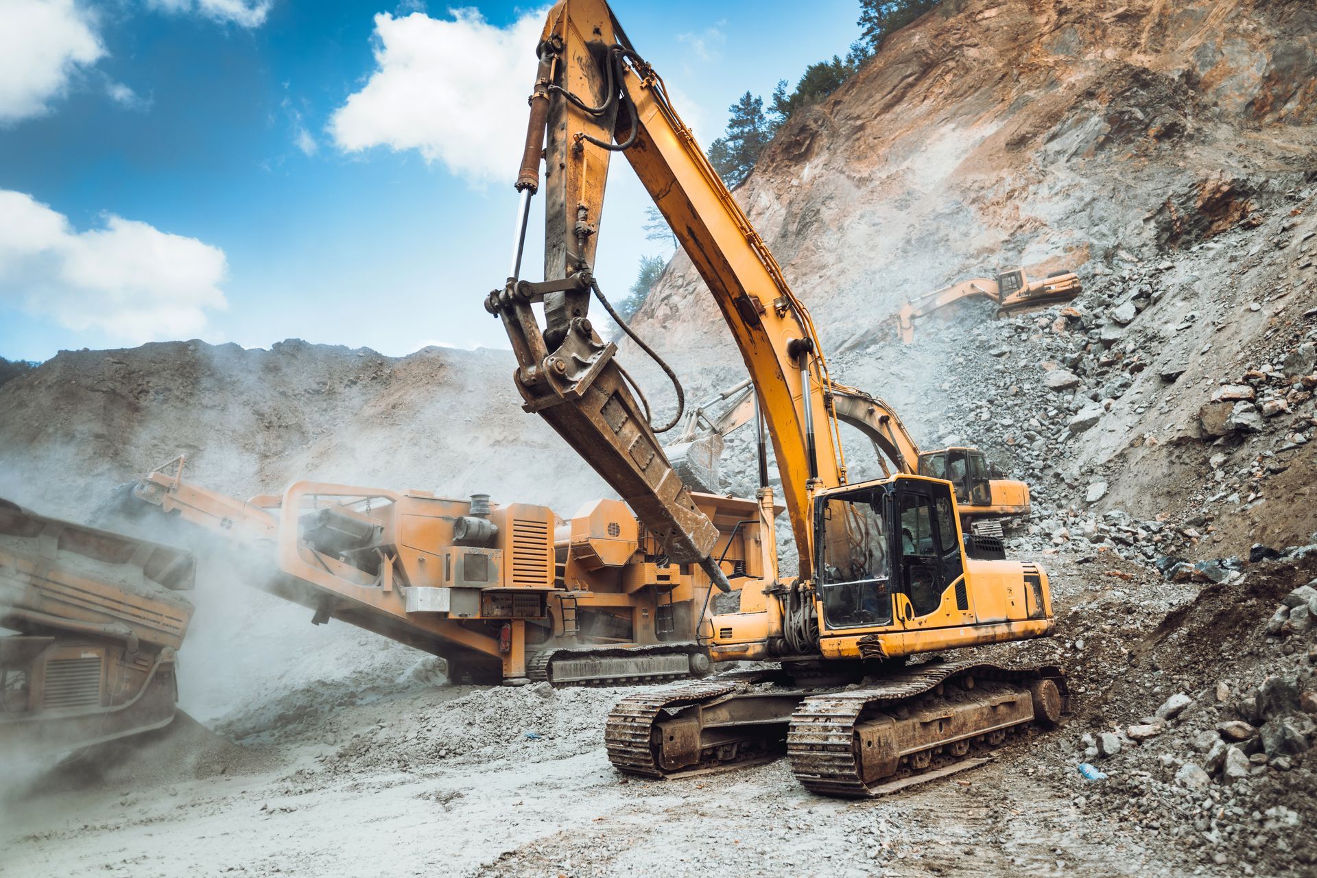 Excavator at a dusty quarry with a dump truck and rocky cliff under a blue sky