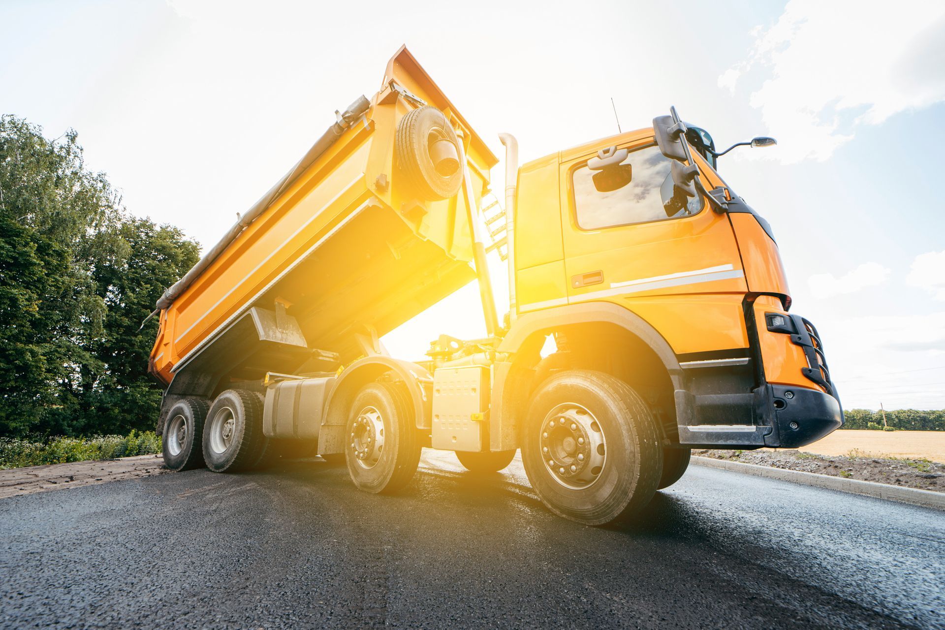 Yellow dump truck on a sunlit road, viewed from the side with bright glare from the sun.