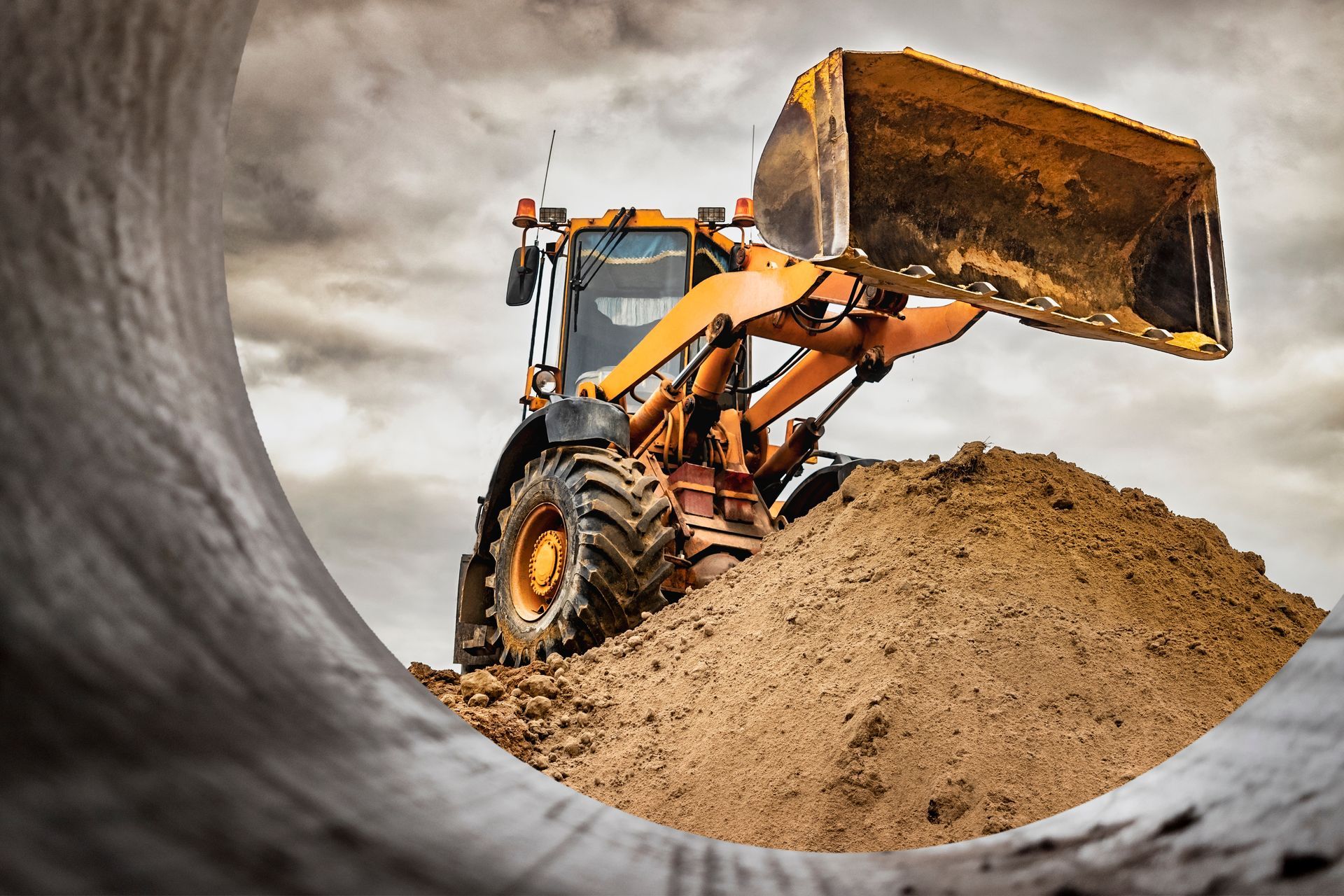 Front loader scooping dirt at a construction site, viewed through a pipe opening