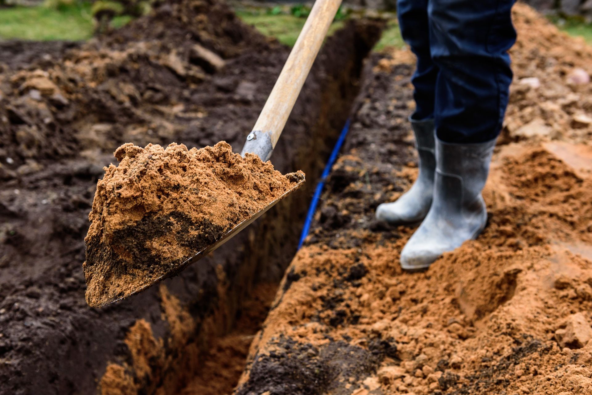 Person digging a trench in muddy soil with a shovel, wearing dark pants and gray boots