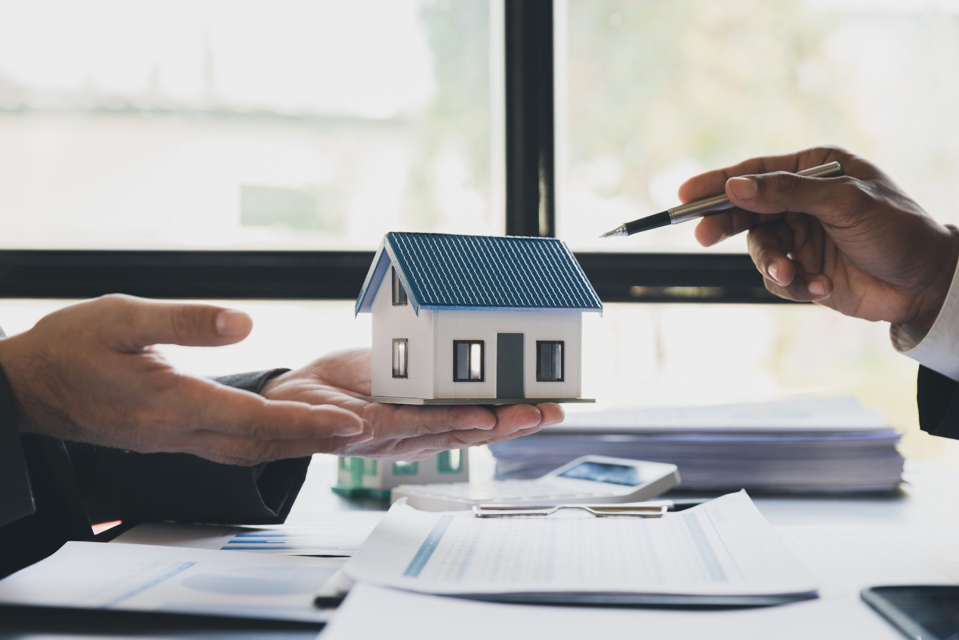 Hands exchanging a small house model over paperwork, suggesting a real estate deal