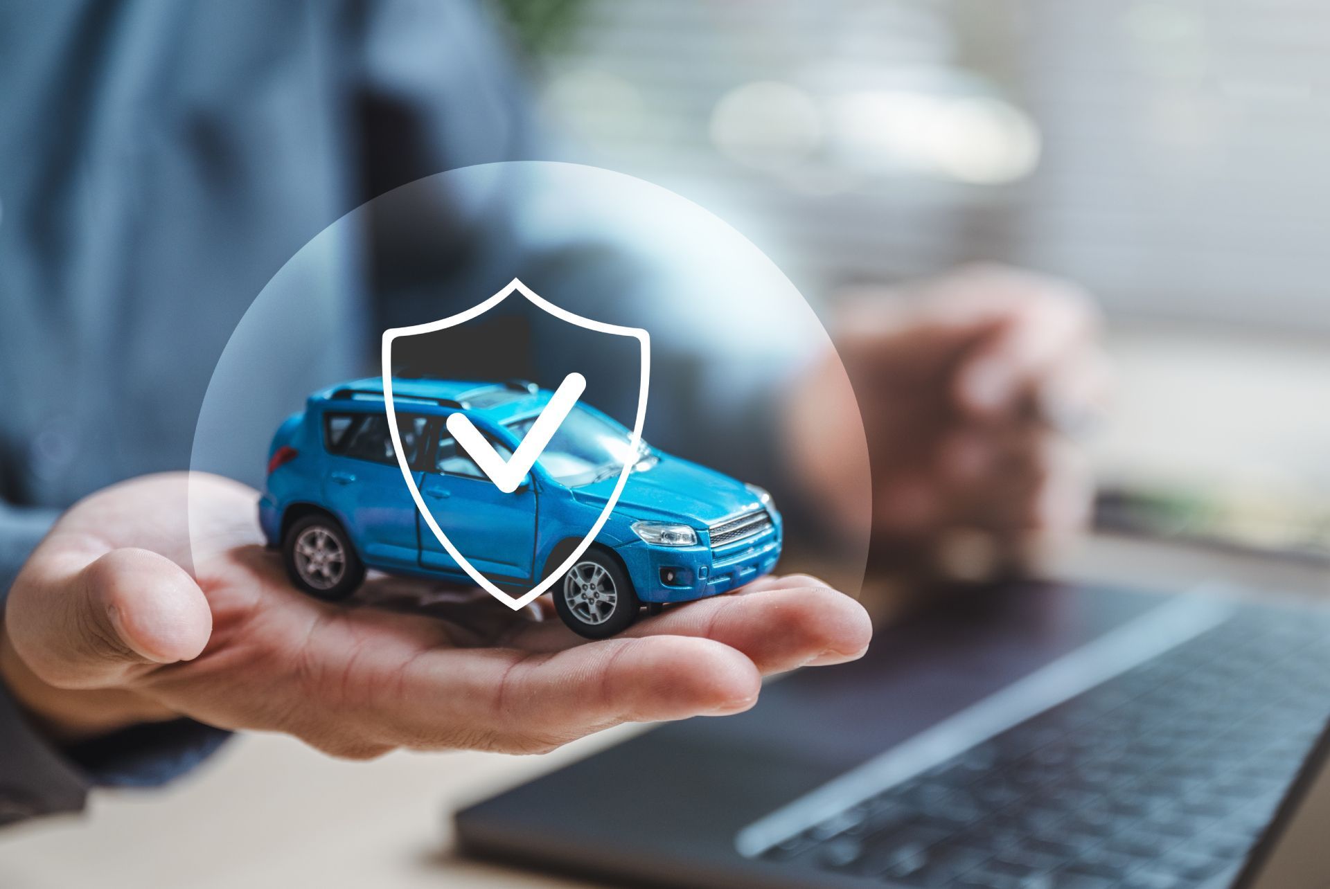 Hand holding a blue toy car under a shield icon, with a laptop in the background
