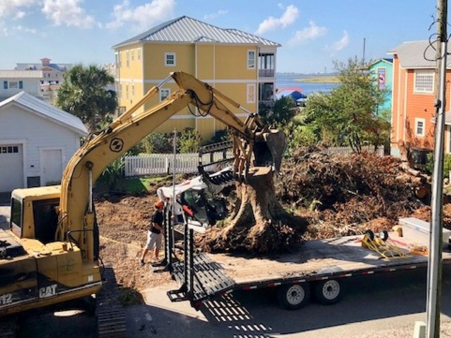 Excavator loads uprooted tree debris onto a trailer in a residential street.