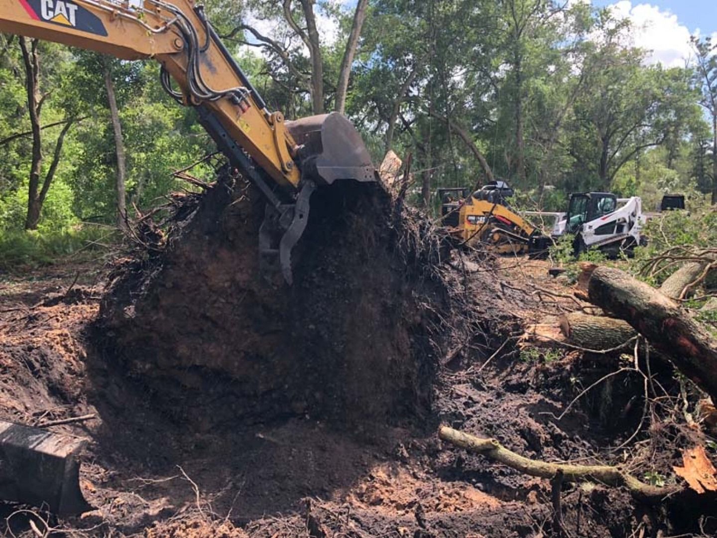 Excavator digging a dirt mound at a wooded construction site, with trucks in the background