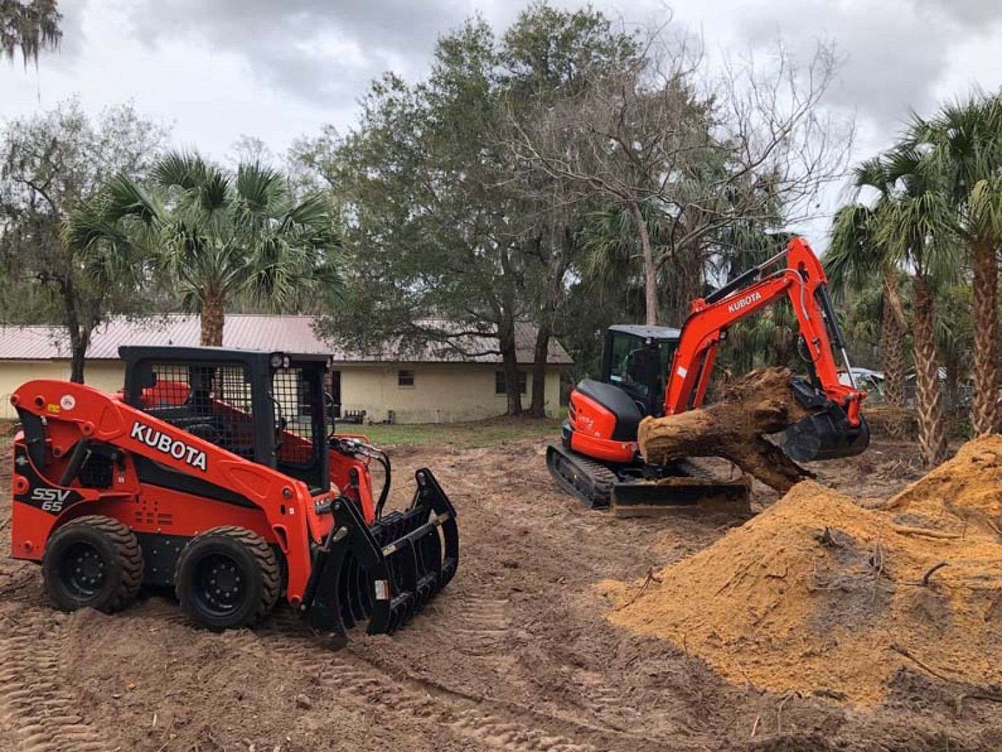 Orange and black skid steer and excavator moving dirt at a construction site with piles of sand and trees.