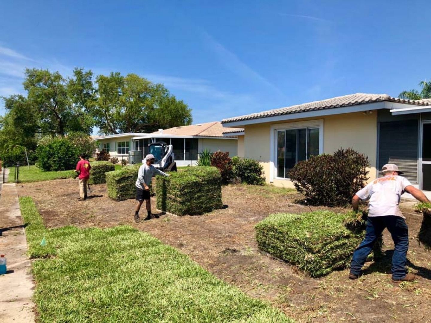 People landscaping a suburban front yard beside a single-story house under a blue sky.
