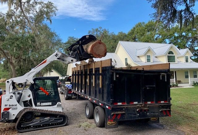 Skid steer loading a large tree log into a black trailer in a suburban driveway