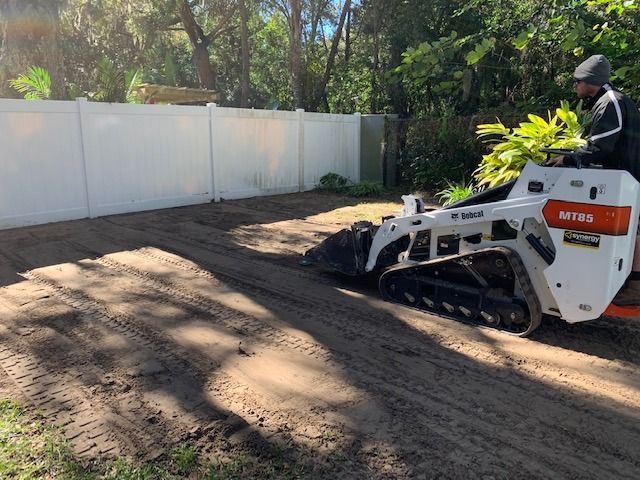 Person operating a white skid steer leveling dirt beside a white fence in a sunny yard