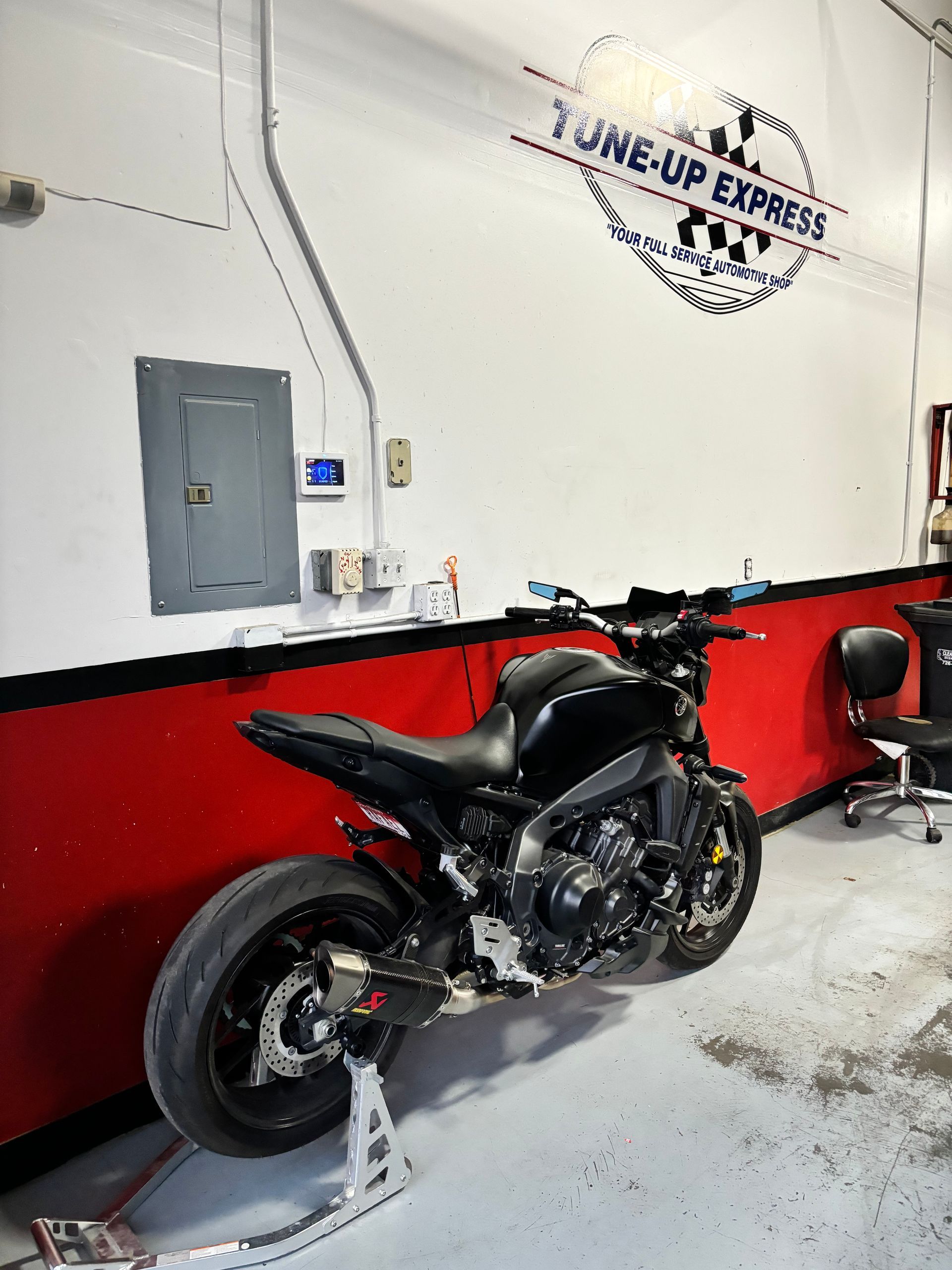 Black motorcycle parked indoors against a white wall with red trim and a logo sign above.  | Tune-Up Express Auto