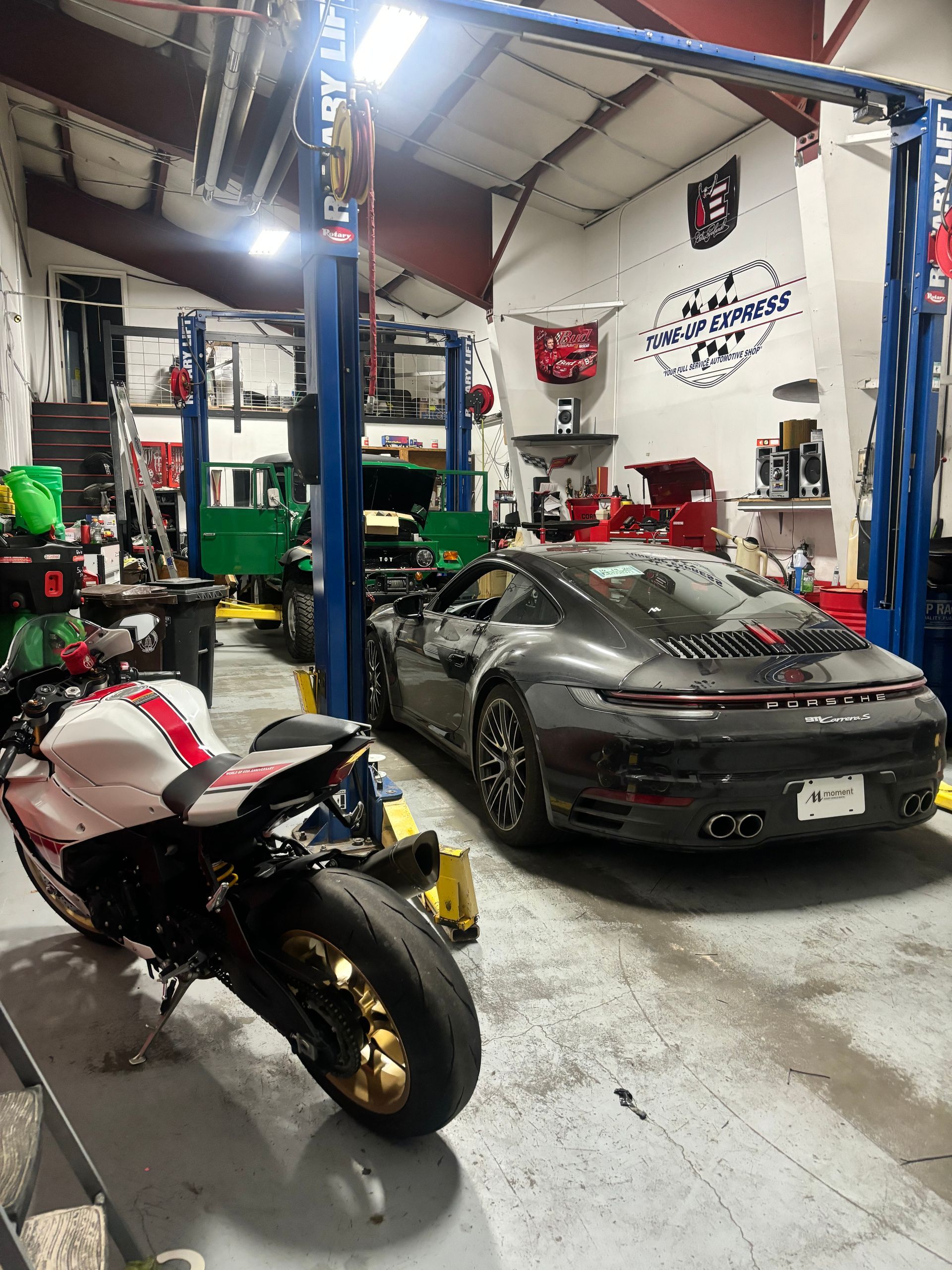 Motorcycles and a sports car inside a cluttered auto repair garage with a lift  | Tune-Up Express Auto