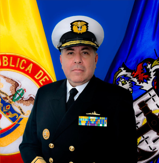 Un oficial naval uniformado con medallas, de pie frente a banderas.