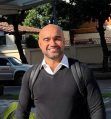 Un hombre con la cabeza rapada sonríe, vistiendo un suéter negro sobre una camisa de cuello blanco
