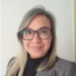 Mujer con gafas y cabello rubio grisáceo sonriendo, vistiendo un blazer gris y un top negro.
