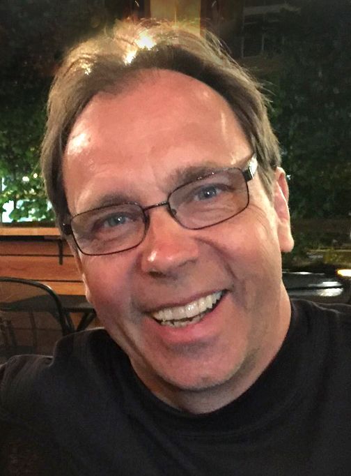 Smiling man with glasses in a dark shirt, seated outdoors at night with warm lights behind him