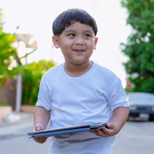 Child holding a tablet outdoors, smiling in a white T-shirt on a sunny street.