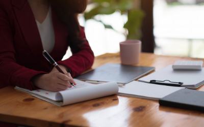 Person writing in a notebook at a wooden desk with a laptop, mug, and tablet nearby