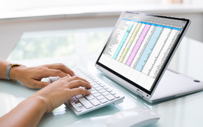 Hands typing on a laptop beside a tablet showing a colorful spreadsheet