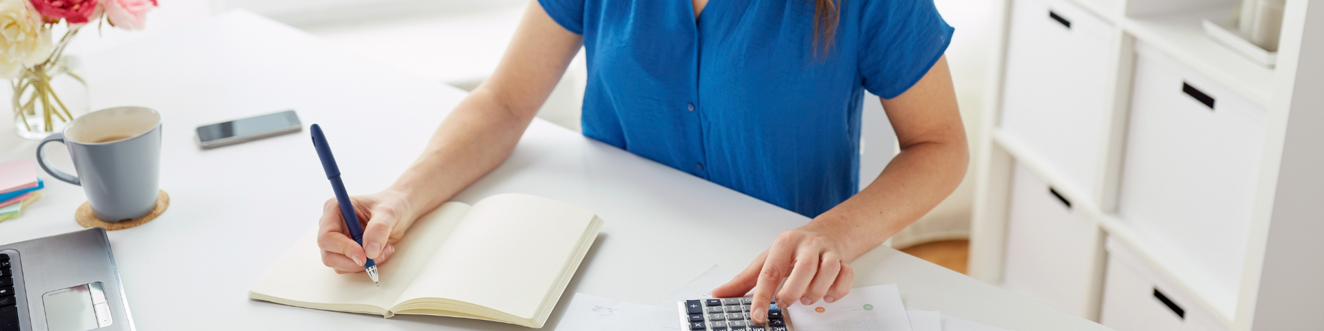 Person writing in an open notebook at a white desk with a blue shirt and a laptop nearby