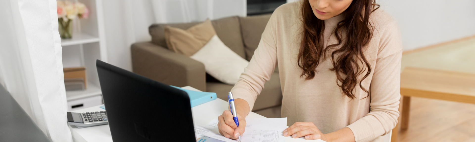 Woman writing beside a laptop at a table in a bright living room