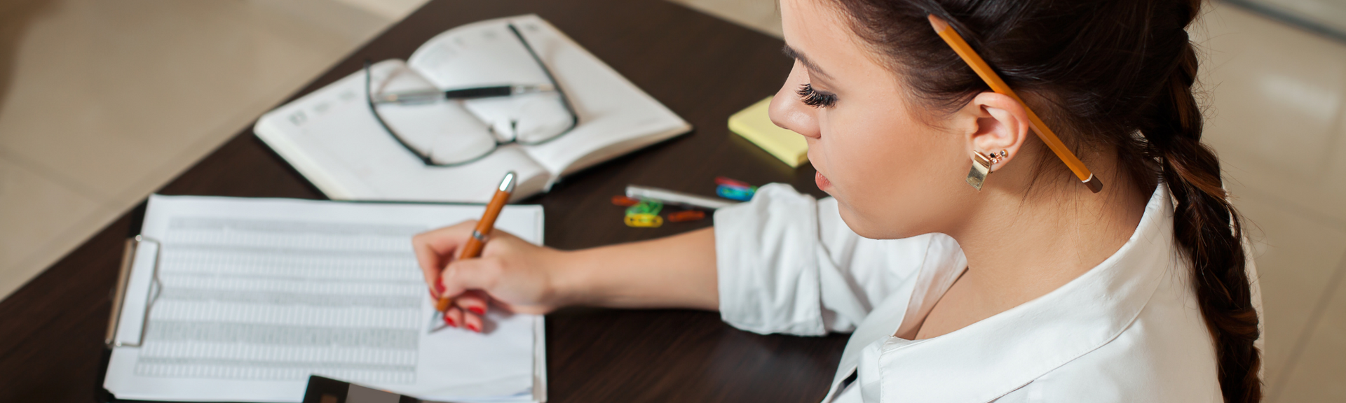 Person writing in a notebook beside a pencil case and eyeglasses on a desk