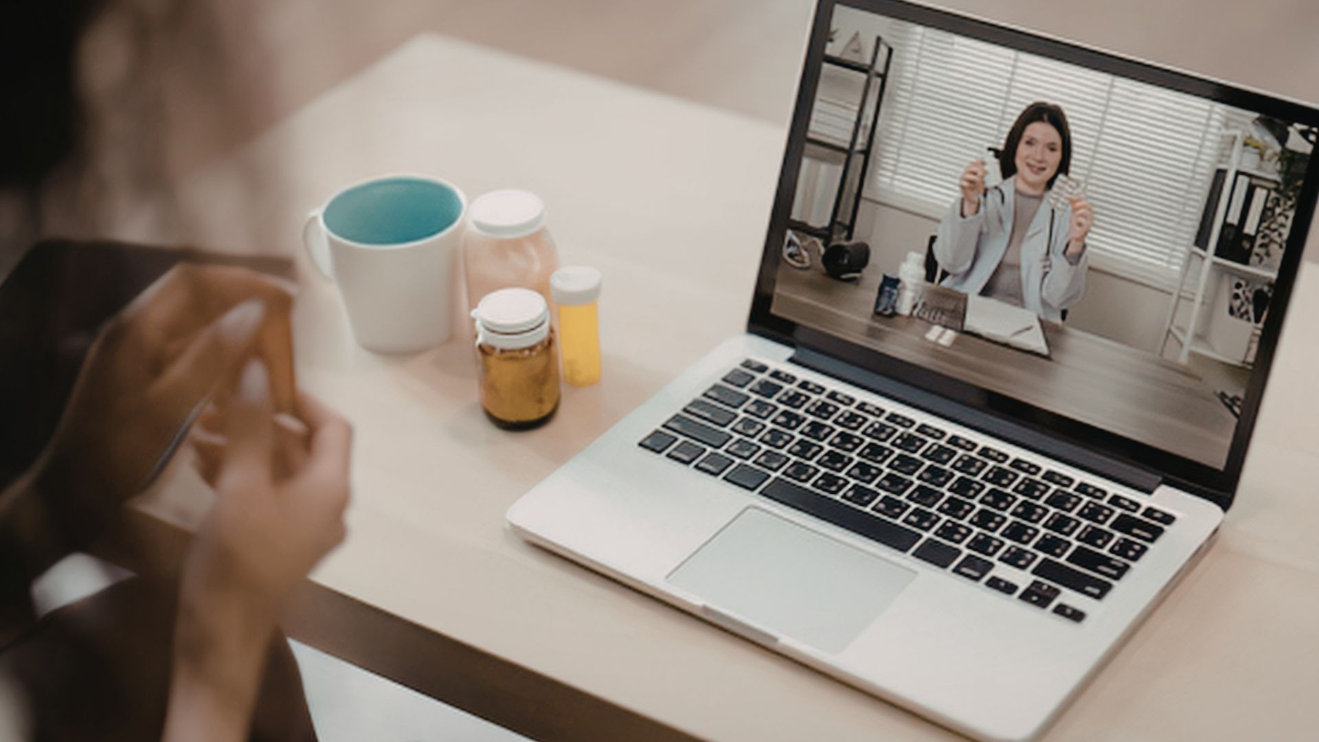 Person on a video call on a laptop beside a mug and pill bottles on a desk