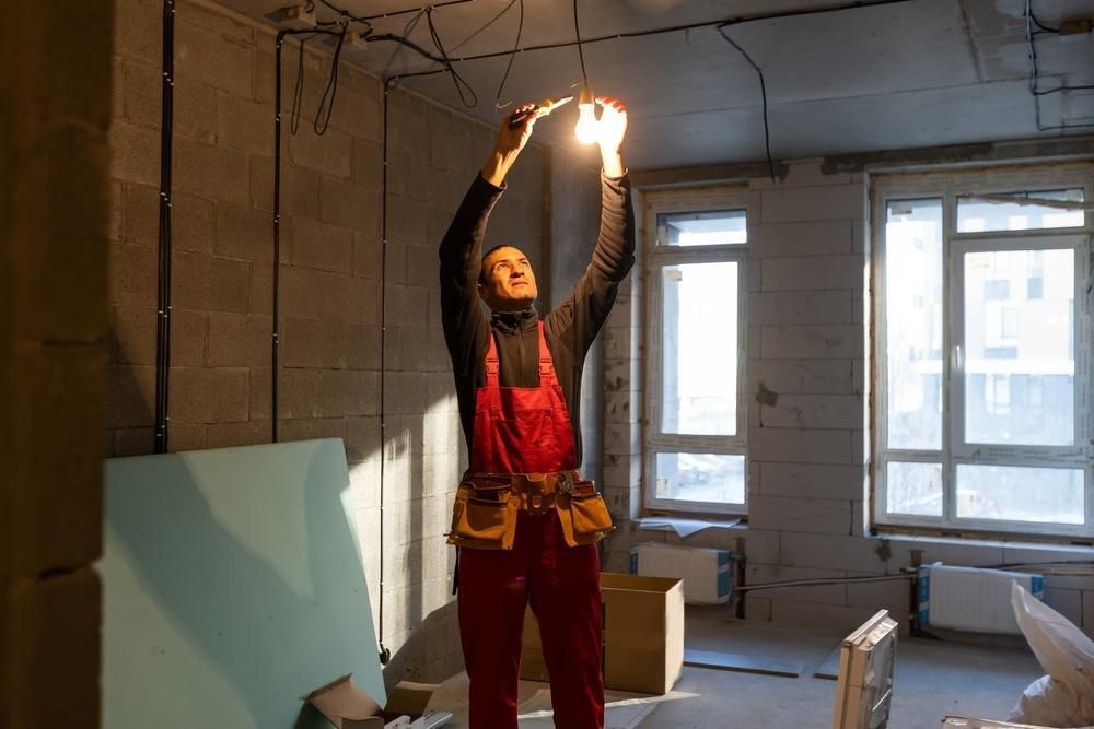 A Man Is Hanging a Light Bulb from The Ceiling in A Room — KARM Electrical Services In Ashtonfield, NSW