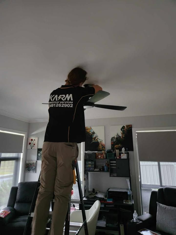 A Man Is Standing on A Ladder Fixing a Ceiling Fan in A Living Room — KARM Electrical Services In Tenambit, NSW