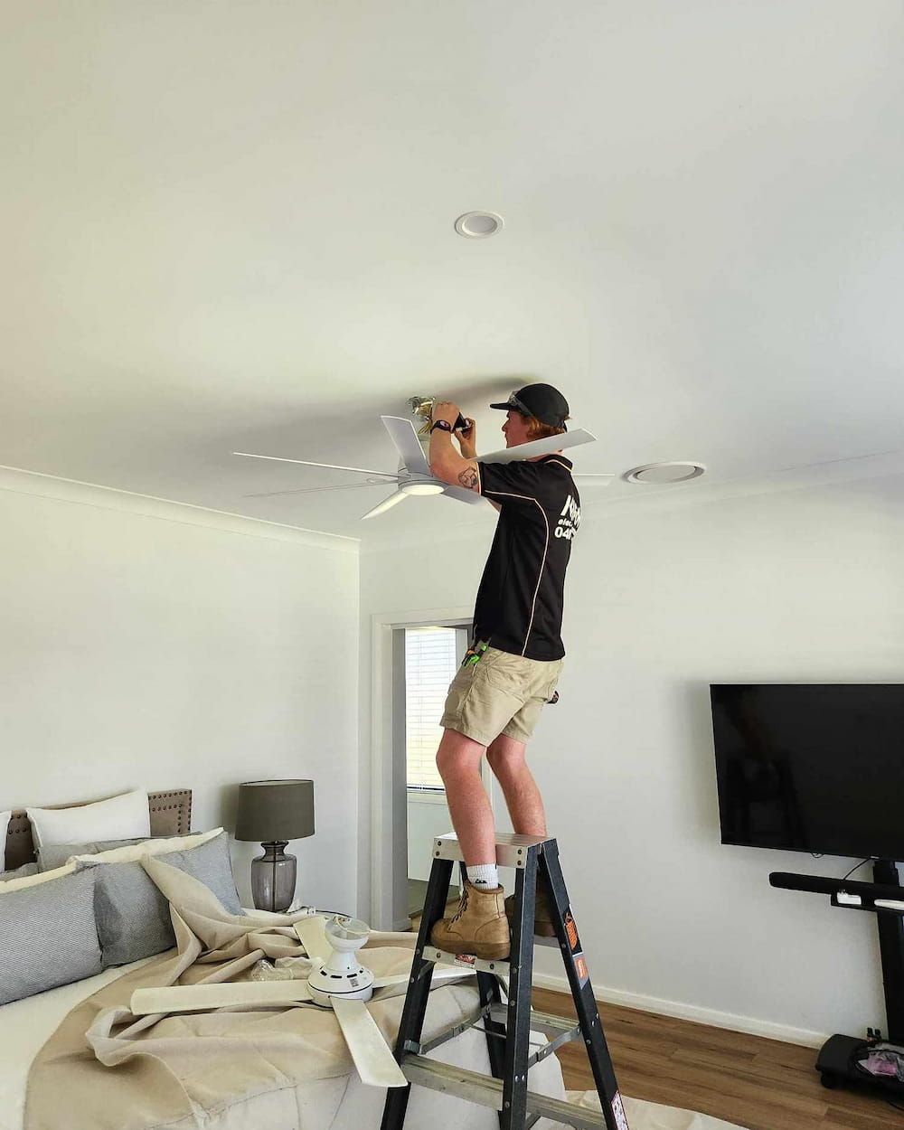A Man Is Standing On A Ladder Fixing A Ceiling Fan — KARM Electrical Services In Raymond Terrace, NSW