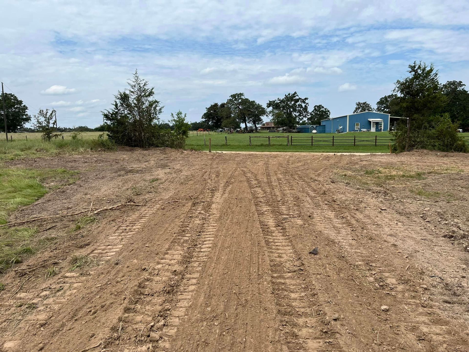 Dirt road with tire tracks, leading to a field with trees and a blue building under a cloudy sky.