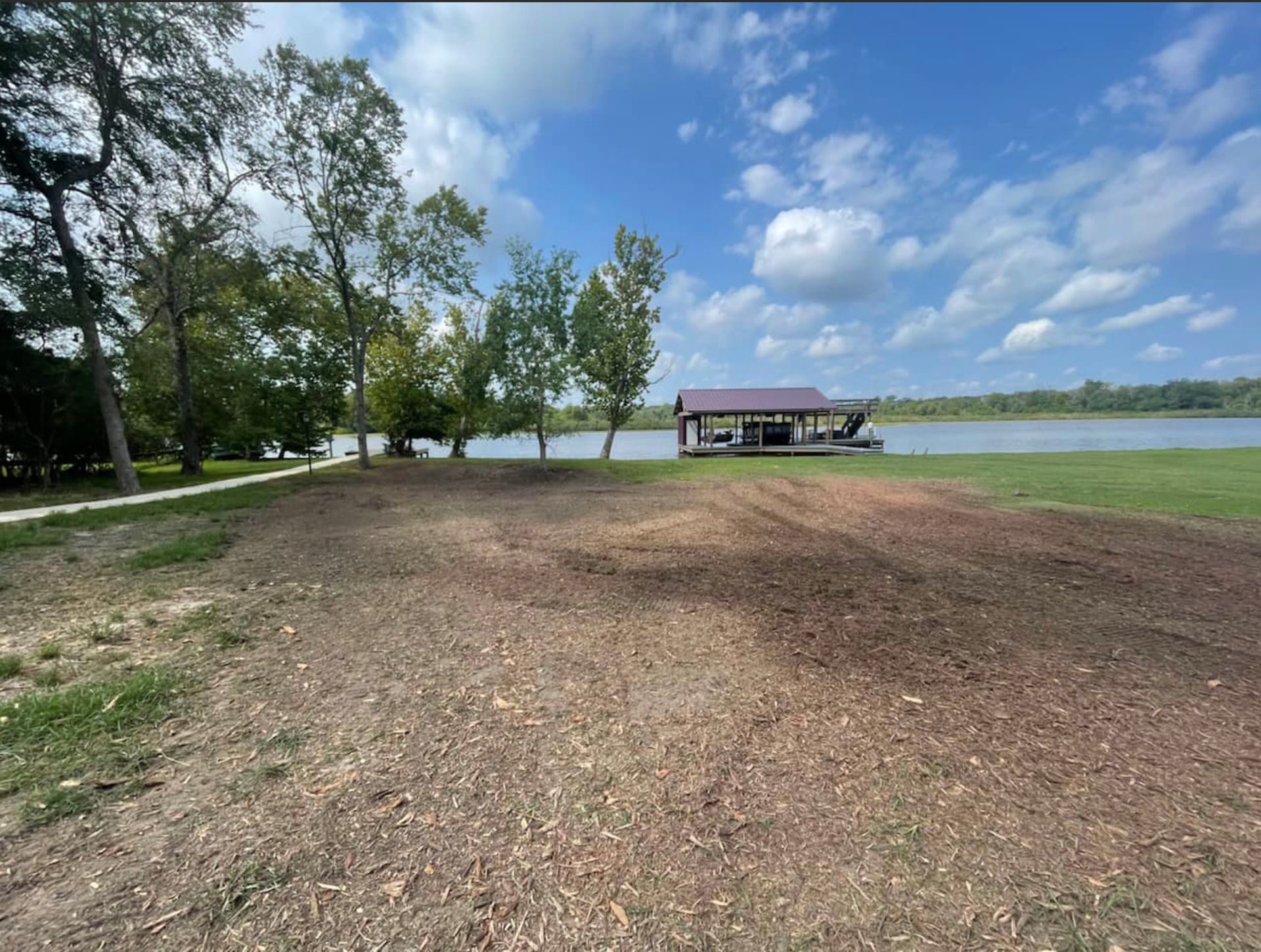 Lawn area near a lake with a covered dock and trees under a blue sky with clouds.