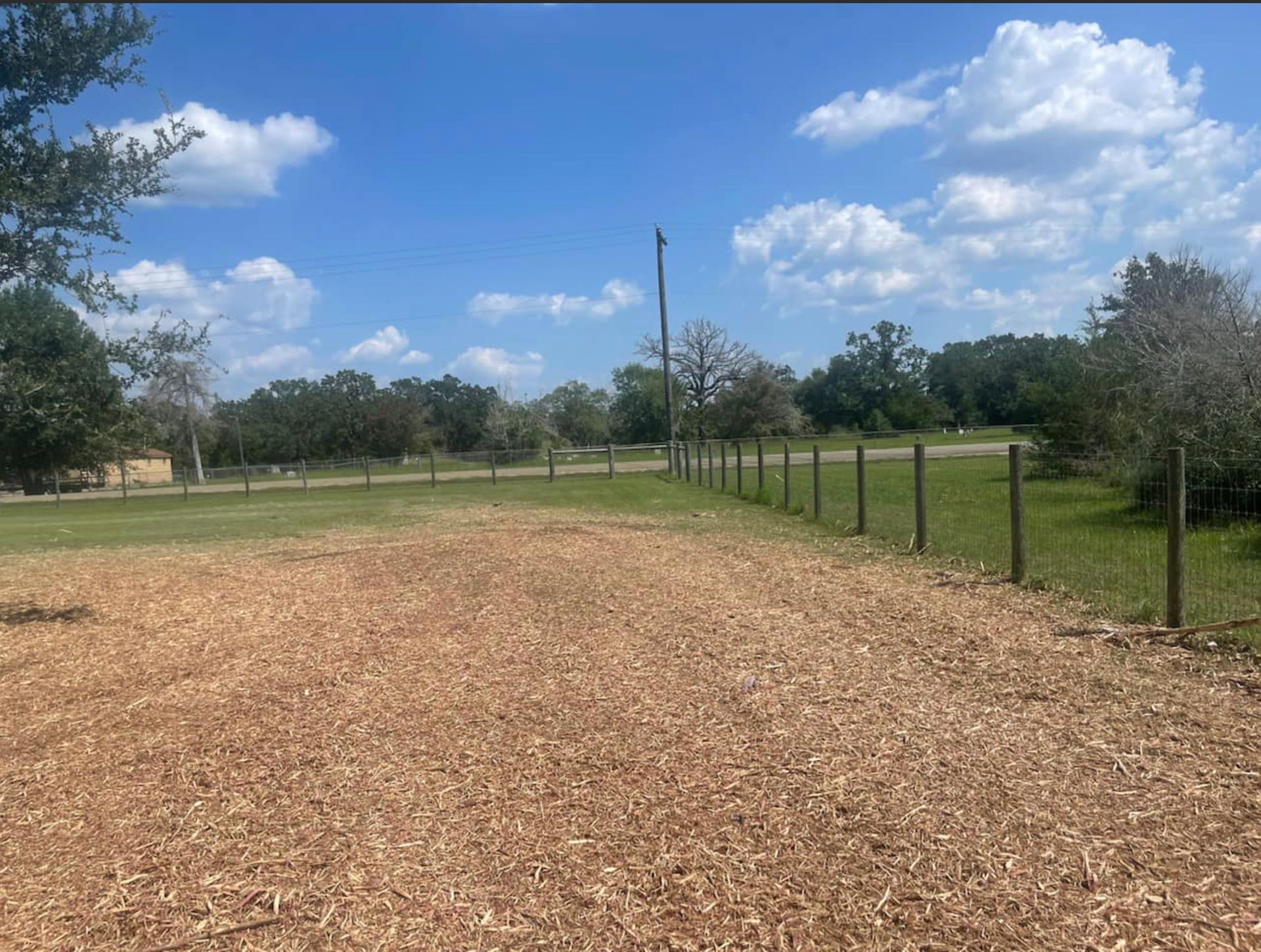 Wood-chipped area with a wooden fence, under a blue sky with fluffy clouds.