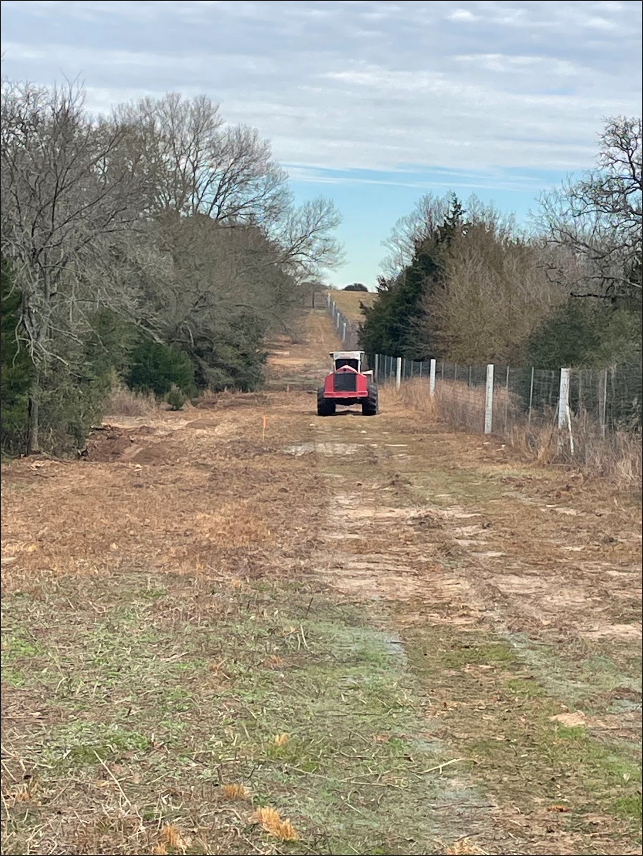 Red tractor driving down a dirt road lined with trees and a fence, under a cloudy sky.