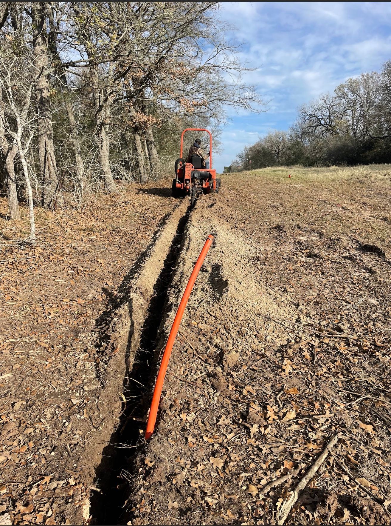 Trench being dug in dry leaves for orange conduit; small tractor in the distance. Trees on either side.