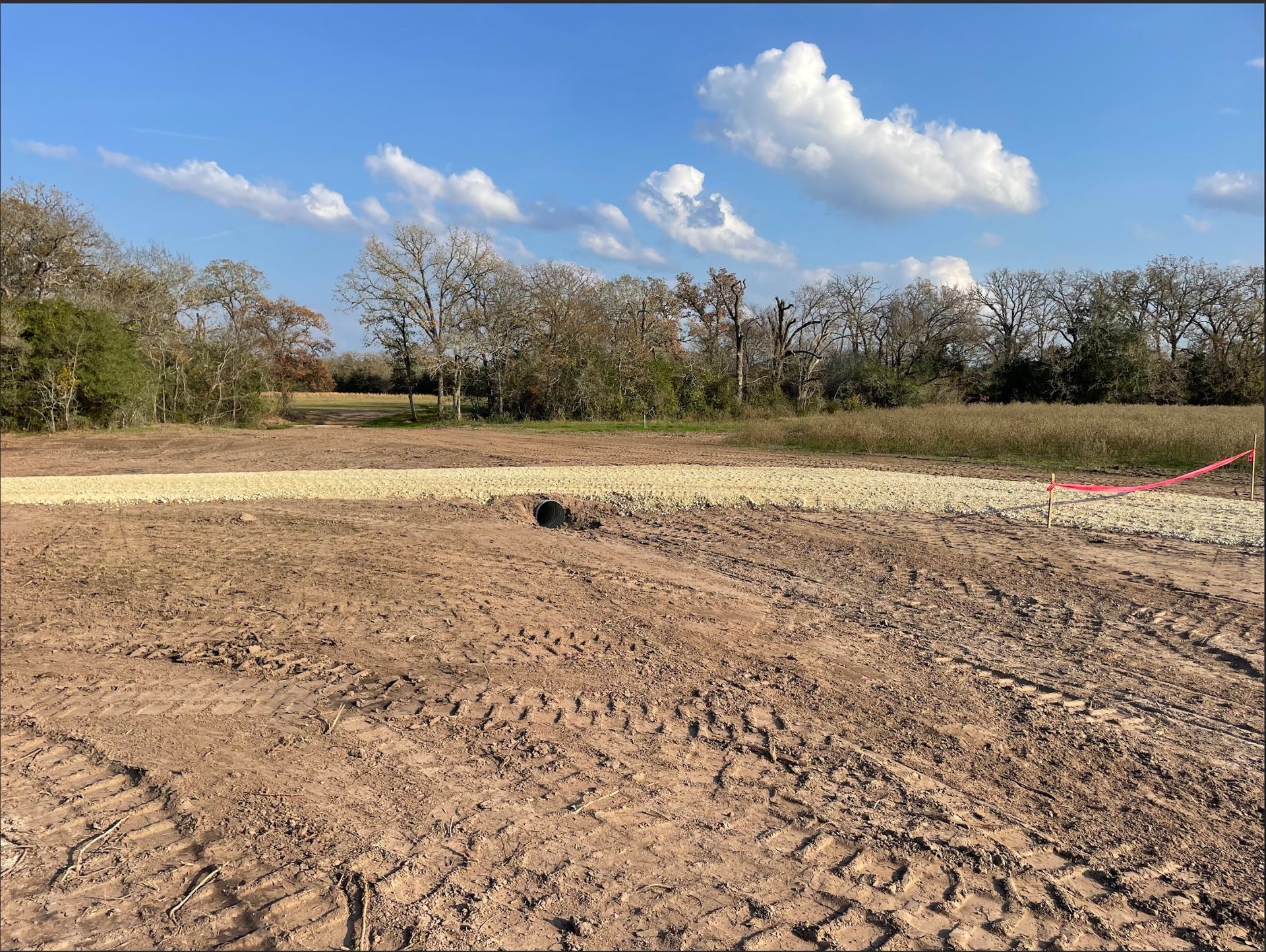Brown, dirt field with tire tracks, gravel border, a small bird, and trees under a blue sky with clouds.