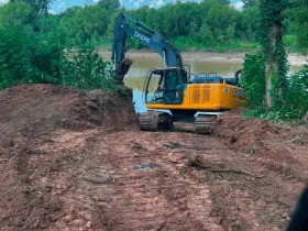 A yellow excavator is digging a hole in the dirt near a river.