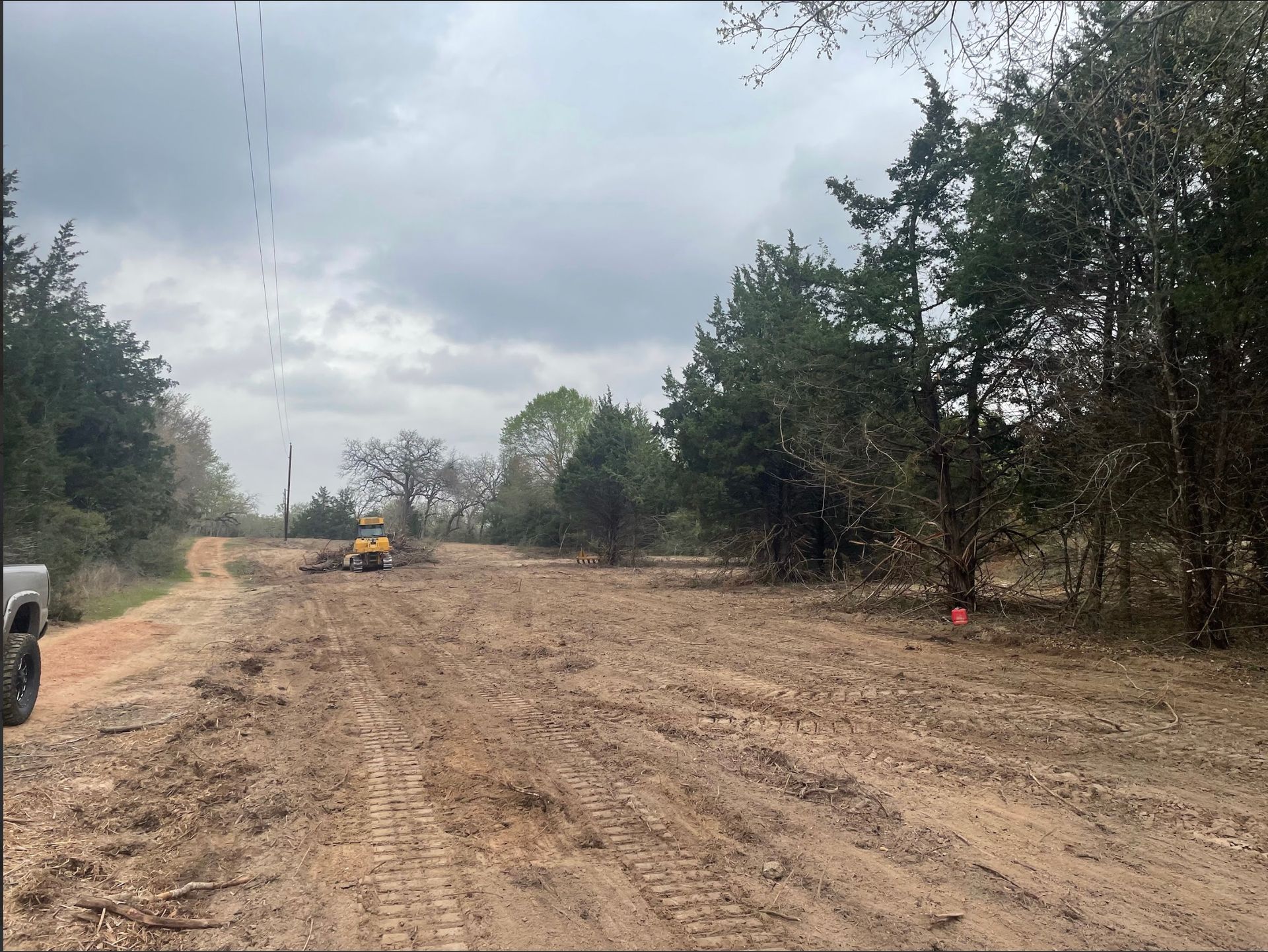 Dirt clearing on a road, excavator in the distance, trees on either side under cloudy skies.