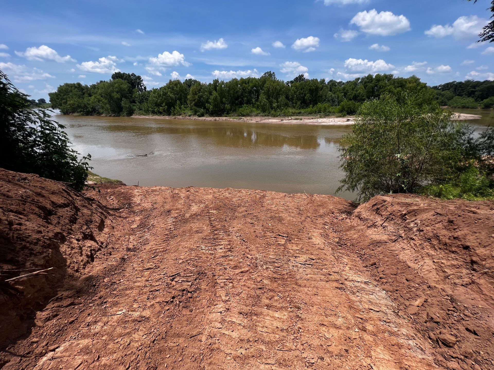 Muddy river view from a dirt embankment under a partly cloudy sky with trees along the river bank.