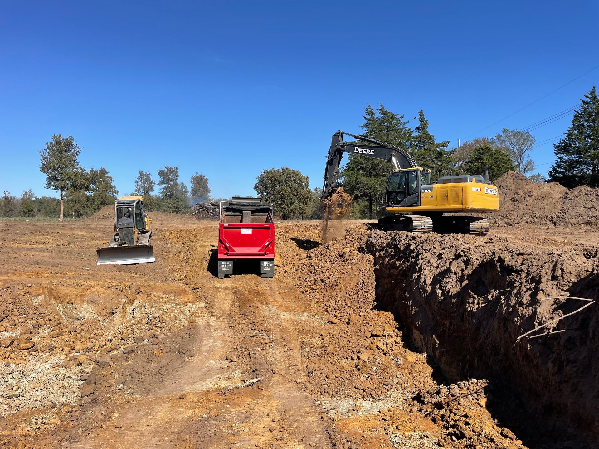 Construction site with excavator, dump truck, and skid steer in a dirt pit under a blue sky.