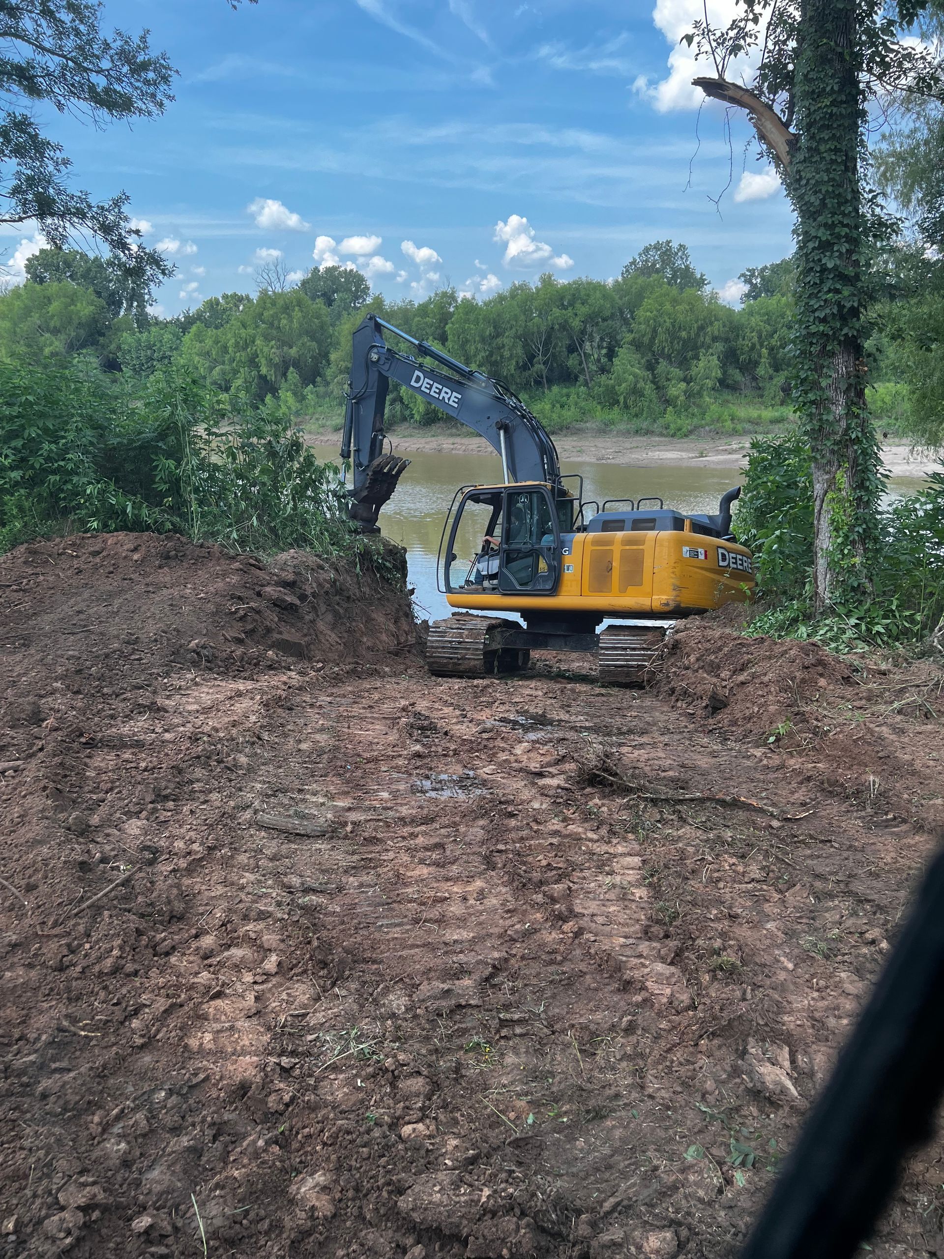 Yellow excavator near a river clearing dirt on a sunny day with blue sky.
