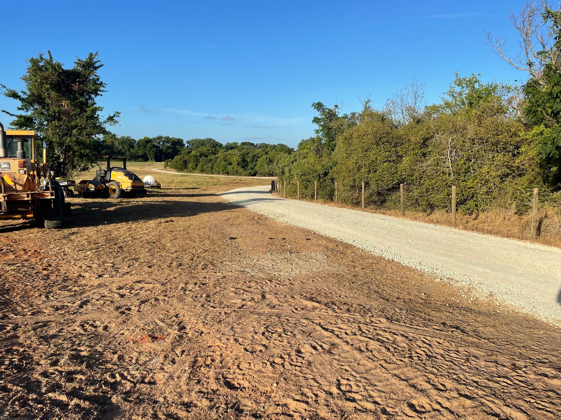 Dirt road under construction with heavy machinery, trees, and clear blue sky.