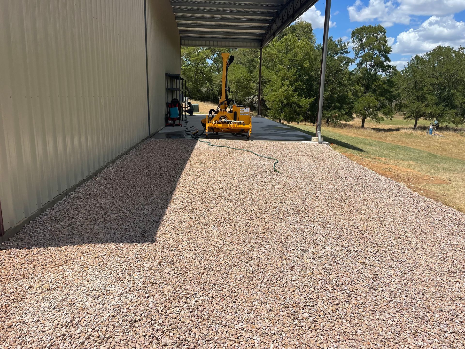A gravel walkway next to a building under a metal awning, with machinery and trees in the background.